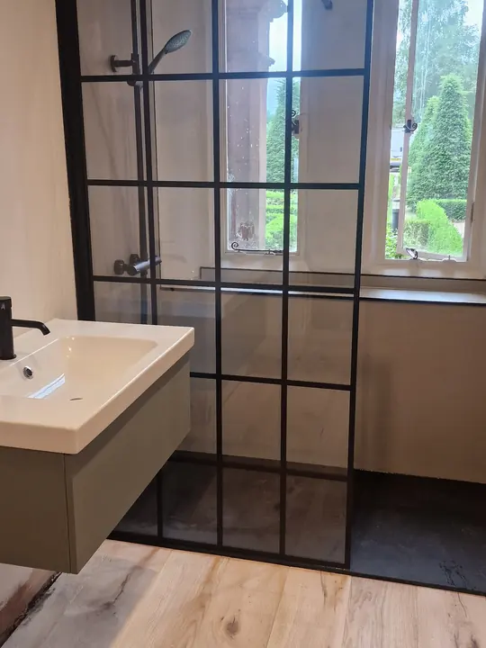 A walk-in shower with a black grid glass screen beside a wall-hung basin and a window.