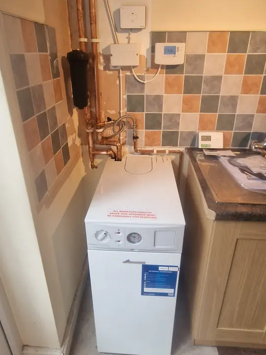 A floor-standing boiler in a utility corner, with copper pipework and heating controls on the tiled wall above.
