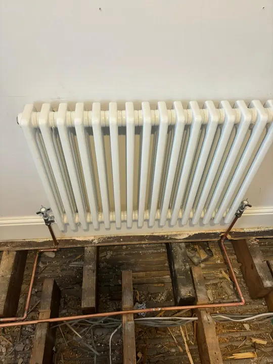 A white column radiator fitted with new copper pipework running beneath the lifted floorboards.