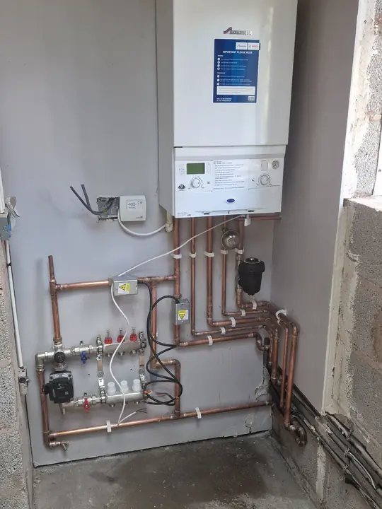A wall-hung boiler with an underfloor heating manifold and copper pipework in a room still being finished.